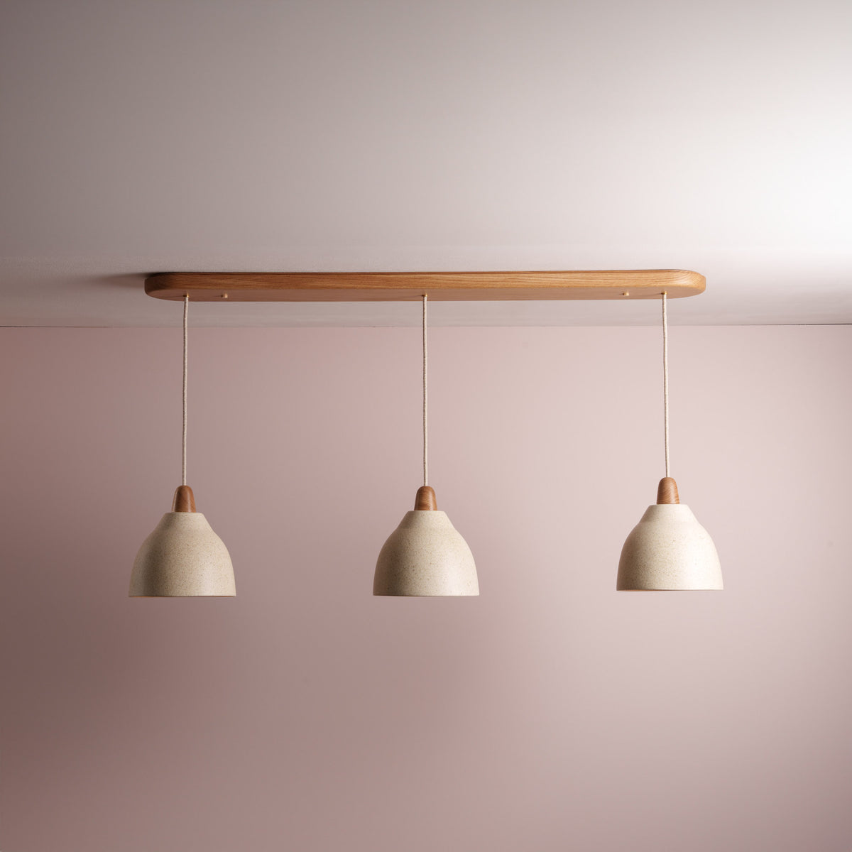 Three cream ceramic pendant lights hanging from an oak wooden bar against a pink wall.